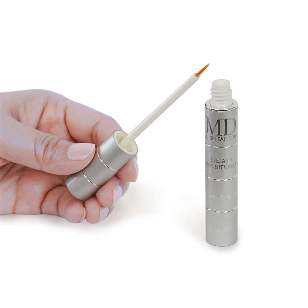 Close-up of hand holding MD Lash Factor 3-Month serum applicator tip next to product showing clear, lightweight formula
